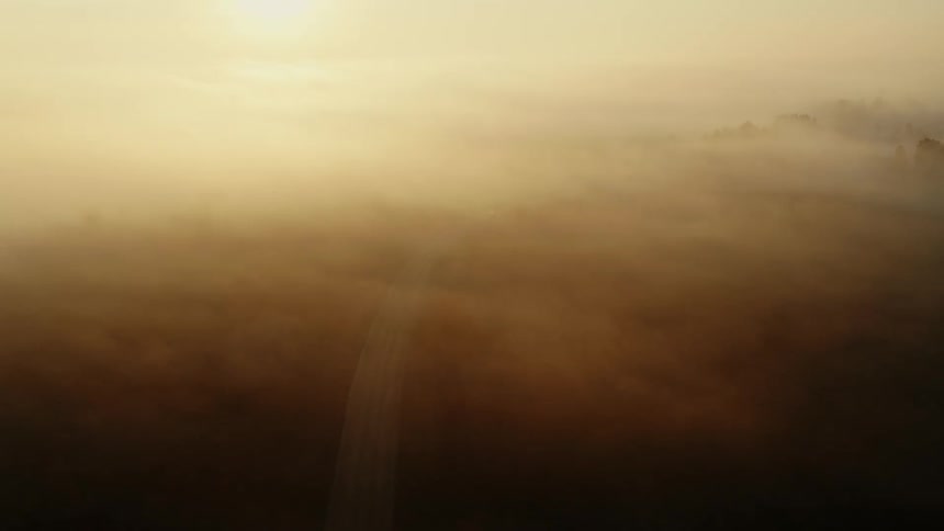 Aerial view of the sun rising in the distance over a highway on a foggy morning with a single car driving into the fog down below
