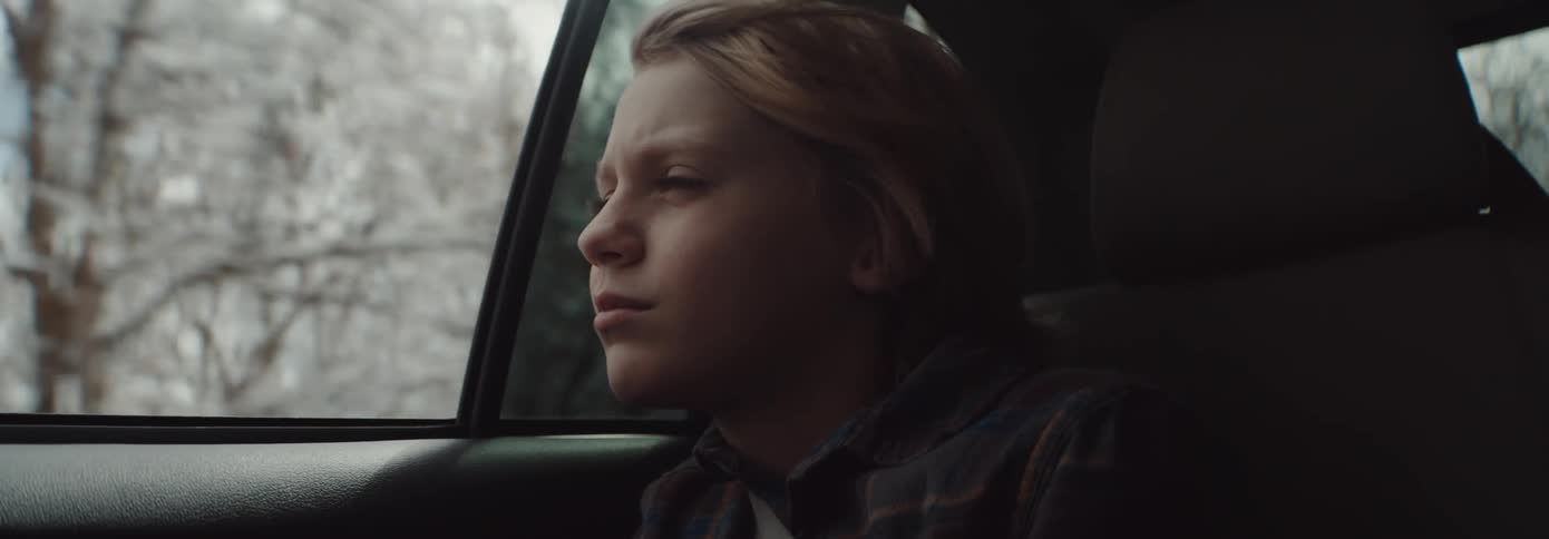 Side view of blonde boy looking out car window of moving car through snowy forest