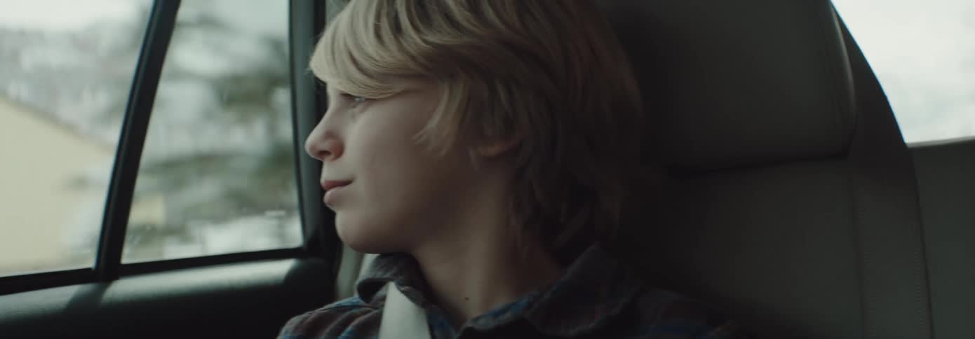 A boy smiles as he rides in the backseat of a vehicle driving past snow covered mountains