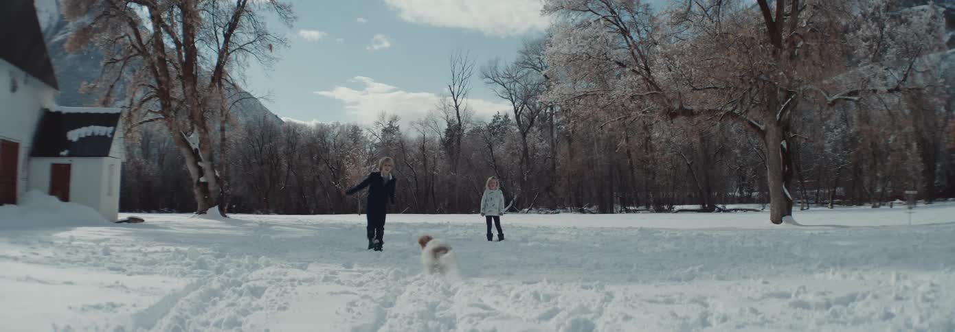 Sister watches brother playing with family dog in snow covered yard with leafless forest and mountains in the background