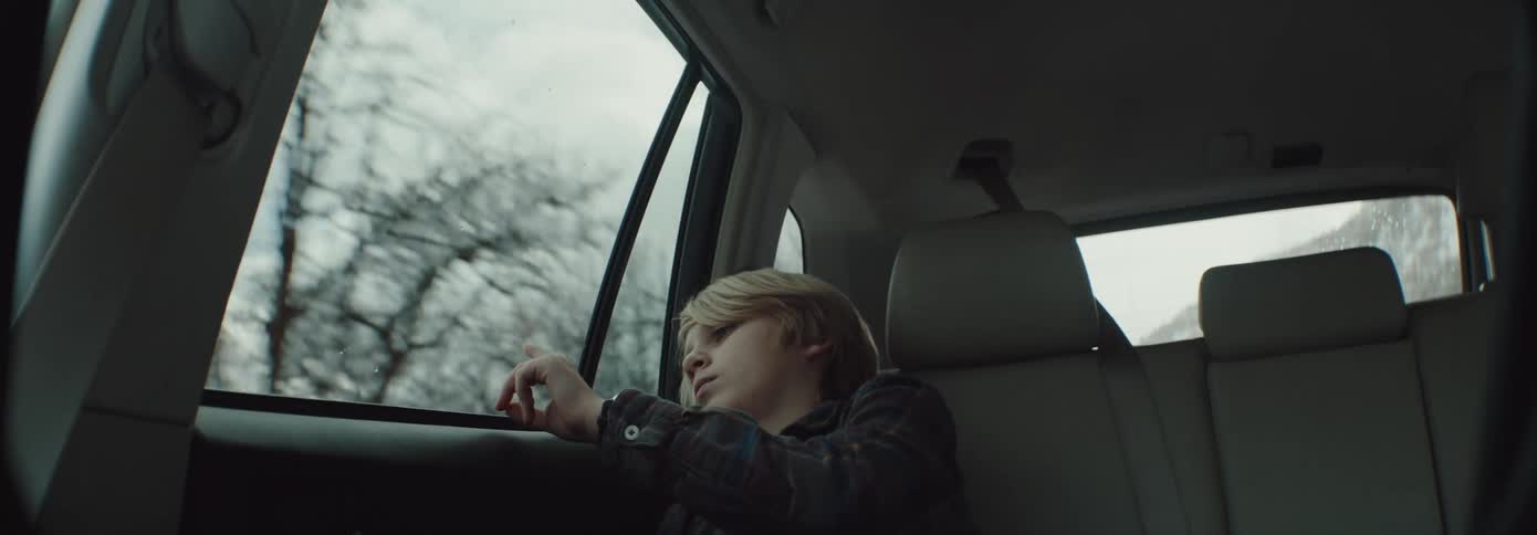 A boy sitting in the back of a car looking bored fiddling with the car window