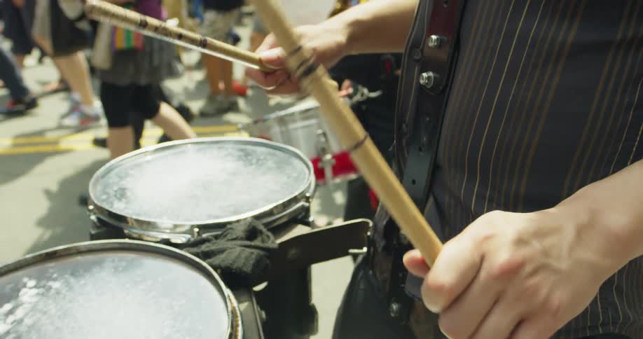 A man drumming on a set of drums hooked around him while walking down a street with a crowd of people
