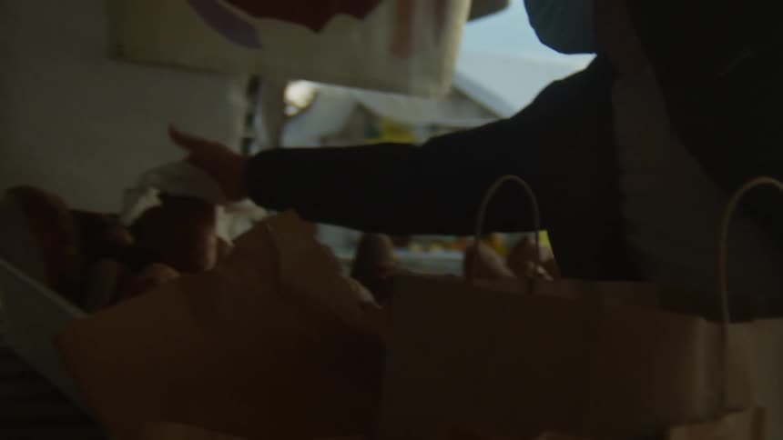 A man retrieves a paper bag and places a loaf of bread inside it in an outdoor market during the day
