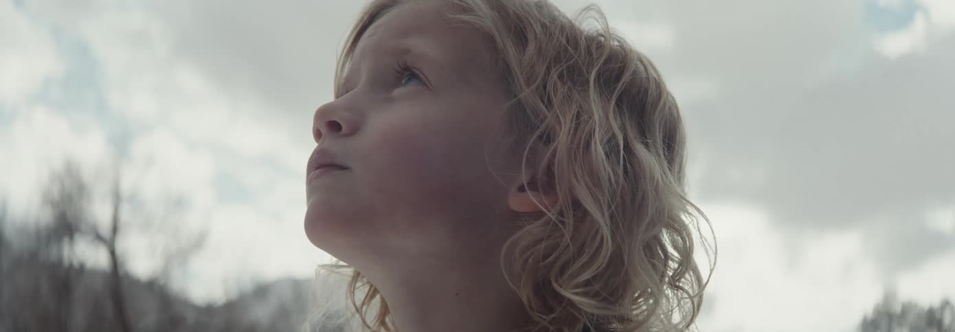 A young girl with curly blonde hair stands outside and looks up toward the sky