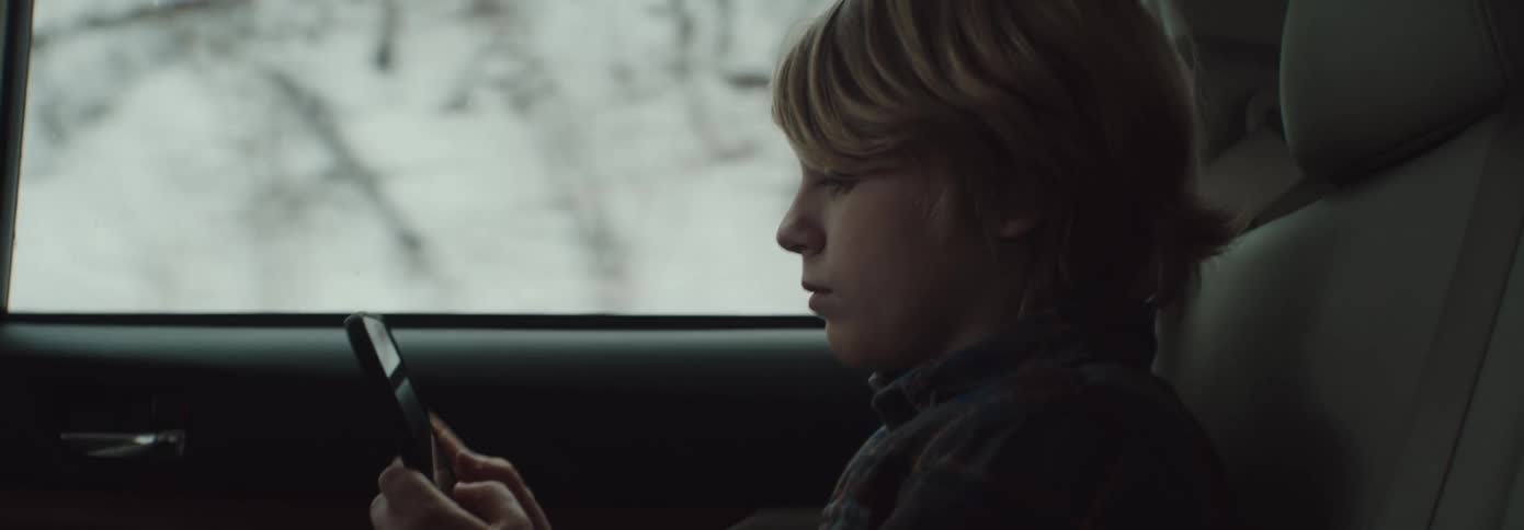 A young boy sits in a moving car driving past snow and trees outdoors while texting on a smart phone