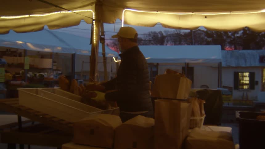 A man under a tent at an outdoor market arranging food in bins