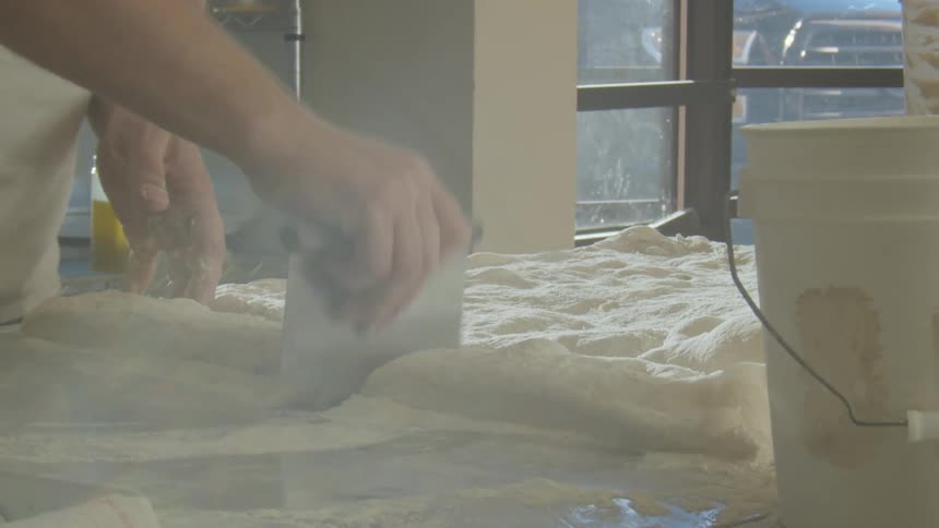 A man cutting and shaping dough on a table inside of a bakery kitchen at daytime