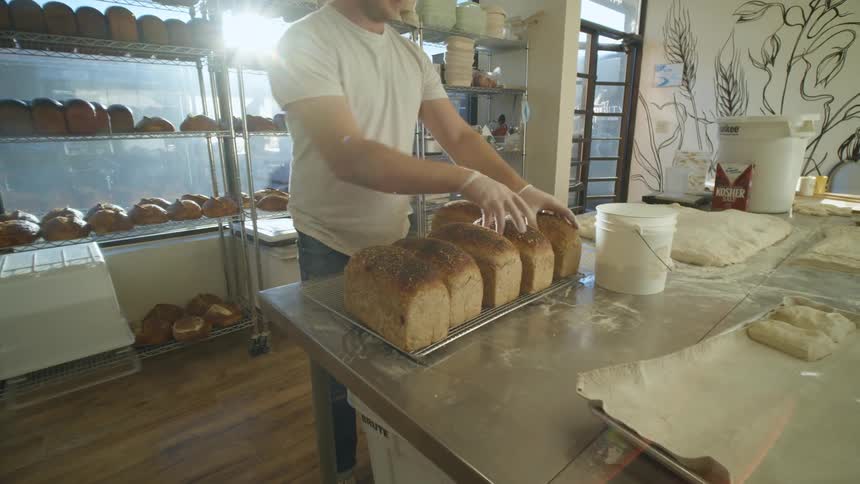 A man in a bakery shop taking bread from a rack and placing it on a table