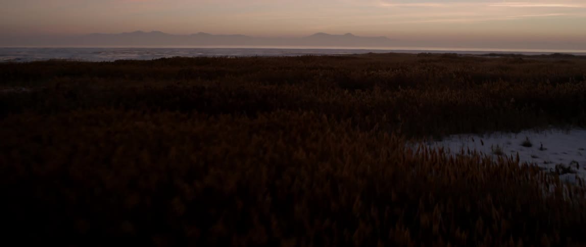 Aerial view of wintry, remote fields and a mountain range in the distance as the sun sets