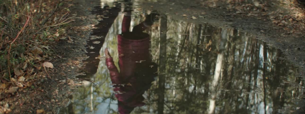 Two women in long puffer coats are reflected in a puddle walking through a forest
