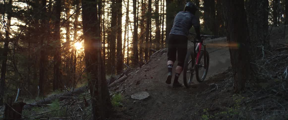 A person with a helmet walking a bike up a dirt path in the forest at sunset