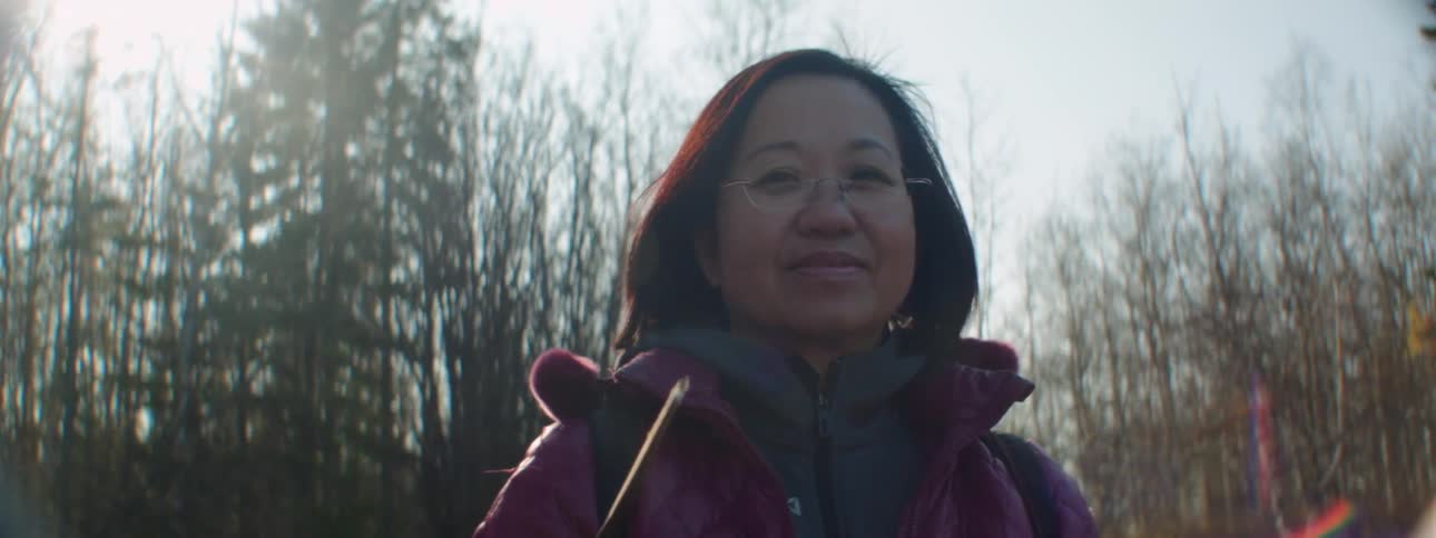 Woman wearing eyeglasses stands smiling next to conifer trees and leafless trees under sunshine