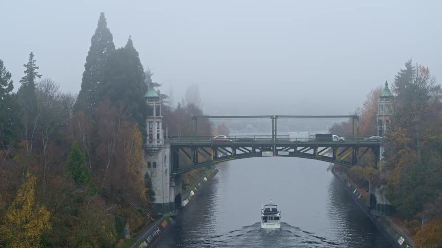 Pullback along the Montlake Cut on a foggy morning as vehicles drive on the bridge and a boat passes underneath