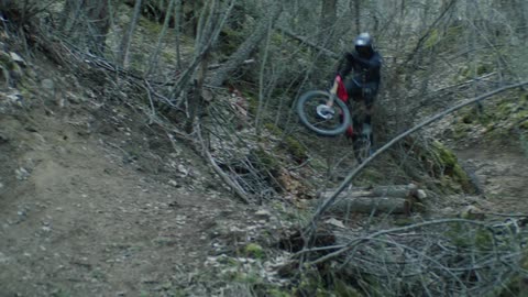 A mountain bike rider jumps in the air as he comes around a curve in a dirt trail filled with fallen tree limbs in the forest