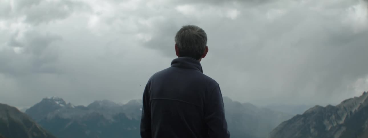 A man looks out at the cloudy sky over a mountain range during the day