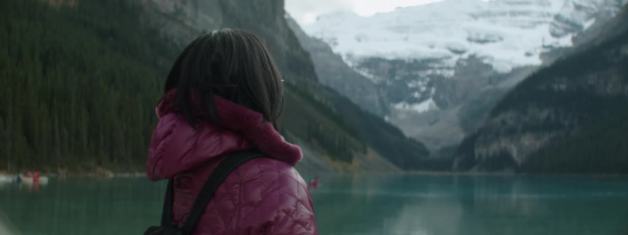Woman wearing hooded coat and backpack stands looking out onto lake and mountains