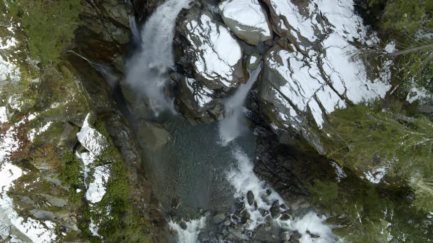 A river flows past snow covered rocks and trees as it leads to a waterfall