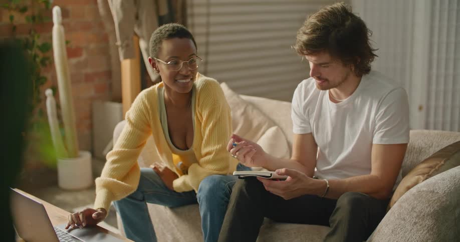 A woman uses a laptop while talking to a man writing in a notebook sitting next to her on a sofa indoors during the day