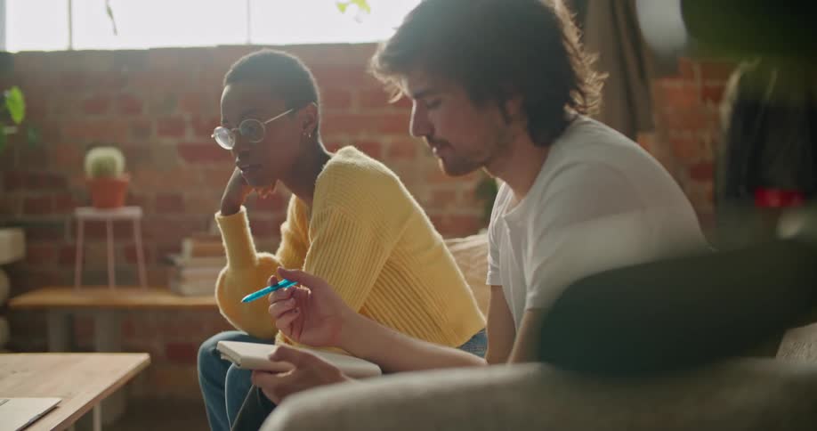 A man holds a notebook and pen sitting beside a woman who points at a laptop on a coffee table