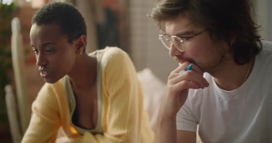A woman uses a laptop while speaking to a man sitting next to her on a sofa in the living room of a house during the day