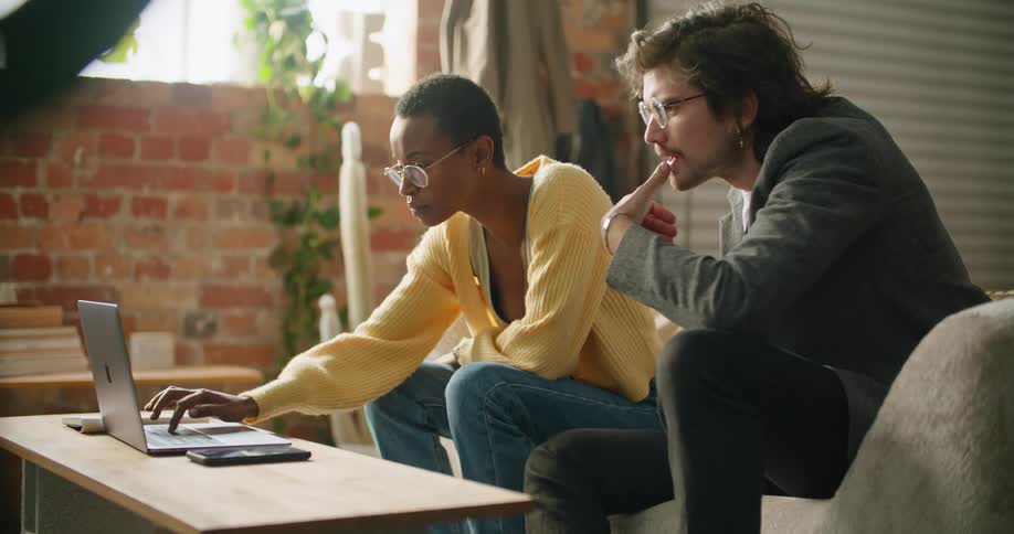 A woman types on a laptop with one hand while speaking with a man sitting next to her on a sofa in a house during the day