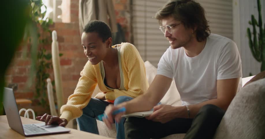 A man and a woman sitting in a room looking at a laptop with a wall and a plant in the background