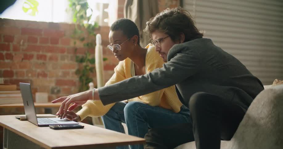 A man touches a laptop screen for emphasis as he watches a friend scroll through while they sit together on the couch