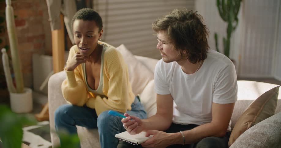A man and woman sit together and the man writes in a small notebook as they talk