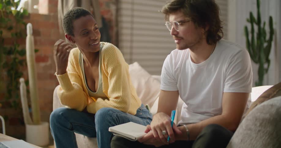 A man and woman sit on a couch and talk and the man writes in a notebook