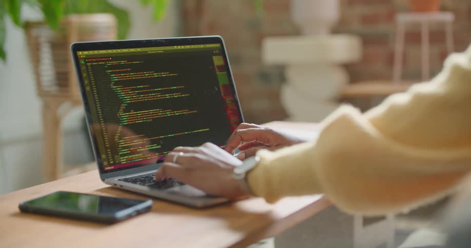 A person in a room sitting by a desk and using a laptop with a wall in the background