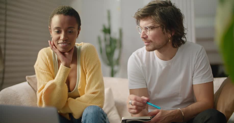 A man and a woman sitting in a room looking at a laptop with a wall and a plant in the background