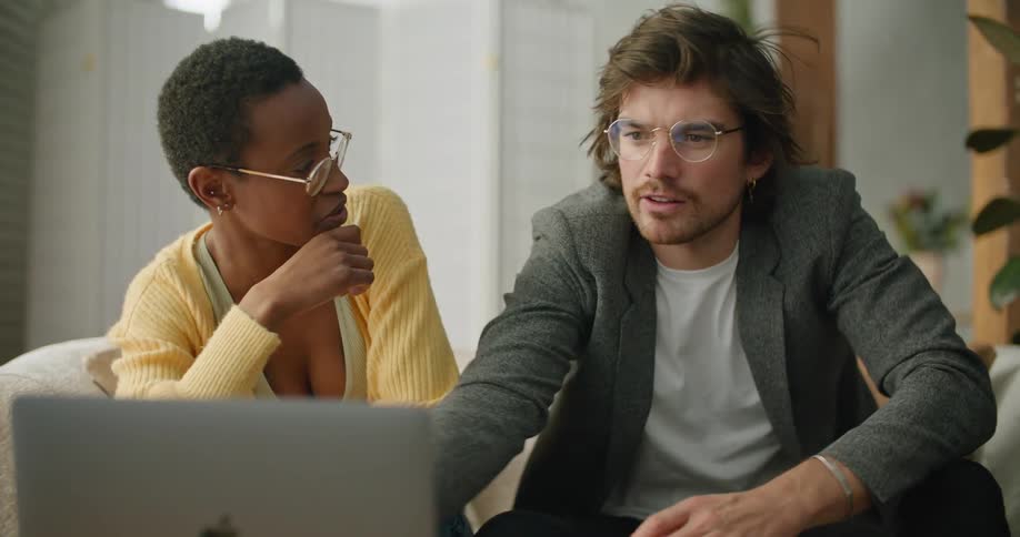 A man and woman sitting side by side on a couch leaning forward and talking while working on a laptop
