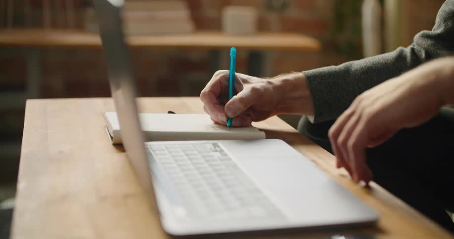 A man chews on his thumb nail makes a note and types on a laptop
