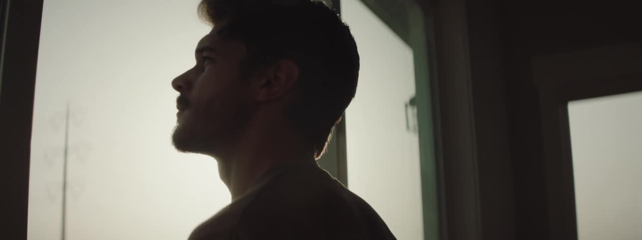 Silhouette of a bearded young man standing in a room looking out a window