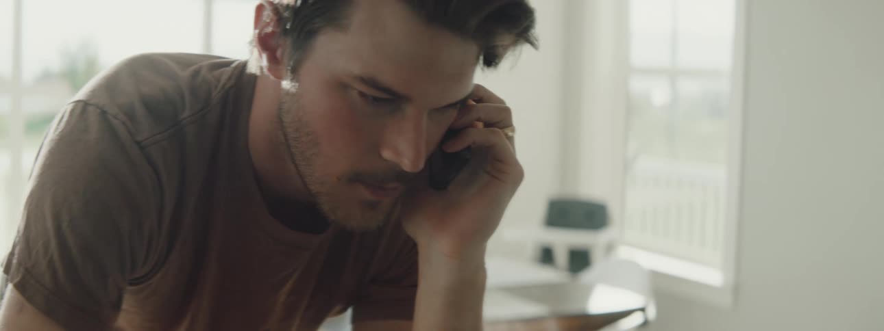 Man types on a laptop while talking on a cellphone indoors