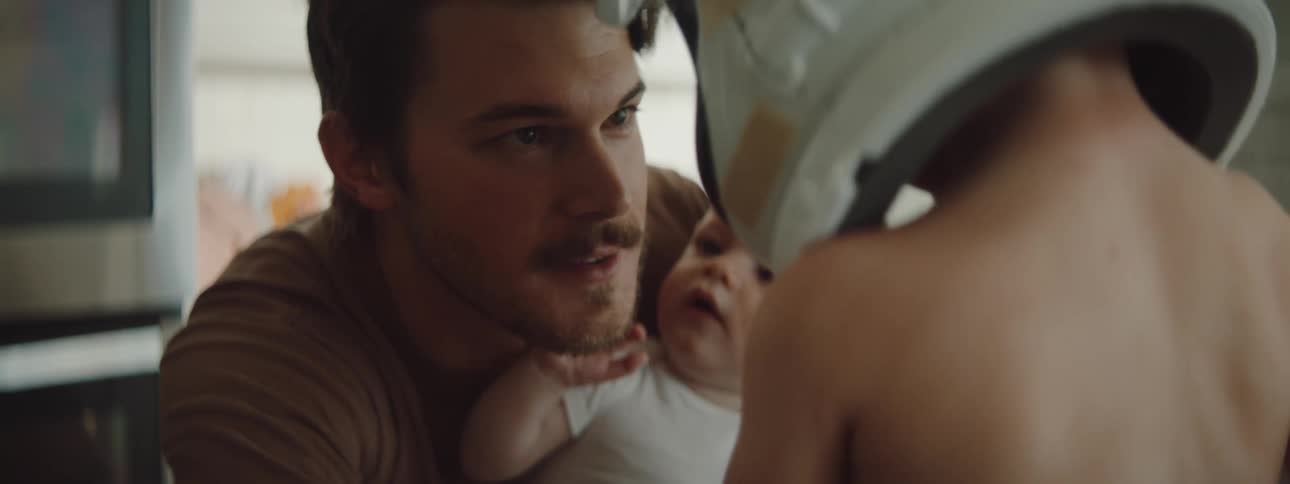 A dad holds his baby while smiling at and talking to his son who is wearing a large helmet