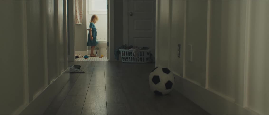 A little girl in the bathroom looking at the toilet with a hallway and a ball in the foreground