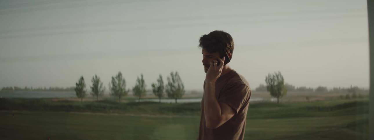 Man with a brown t shirt and a beard talks on the smartphone outdoors