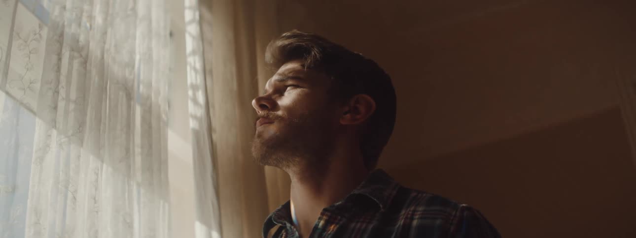A man raises his hand to shield his eyes from the sun as he looks out of a large window in a house during the day