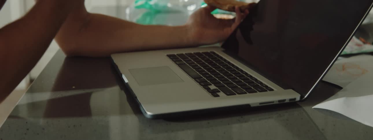 A man is in his kitchen and using a laptop while talking on the phone and eating