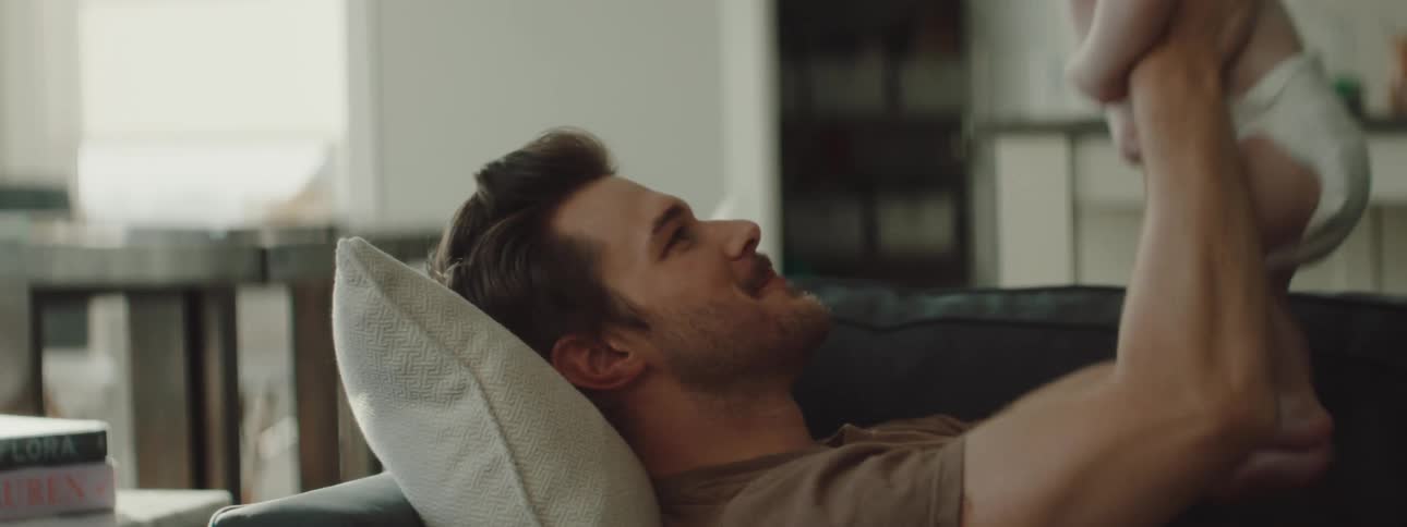 A man laying on a couch playing with a baby with a wall and window in the background