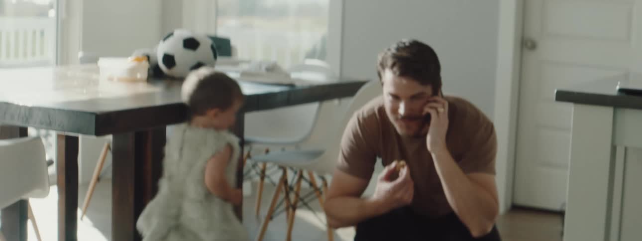 A man eats a snack while talking on the phone and leaning down to greet his kids as they chase each other through the kitchen