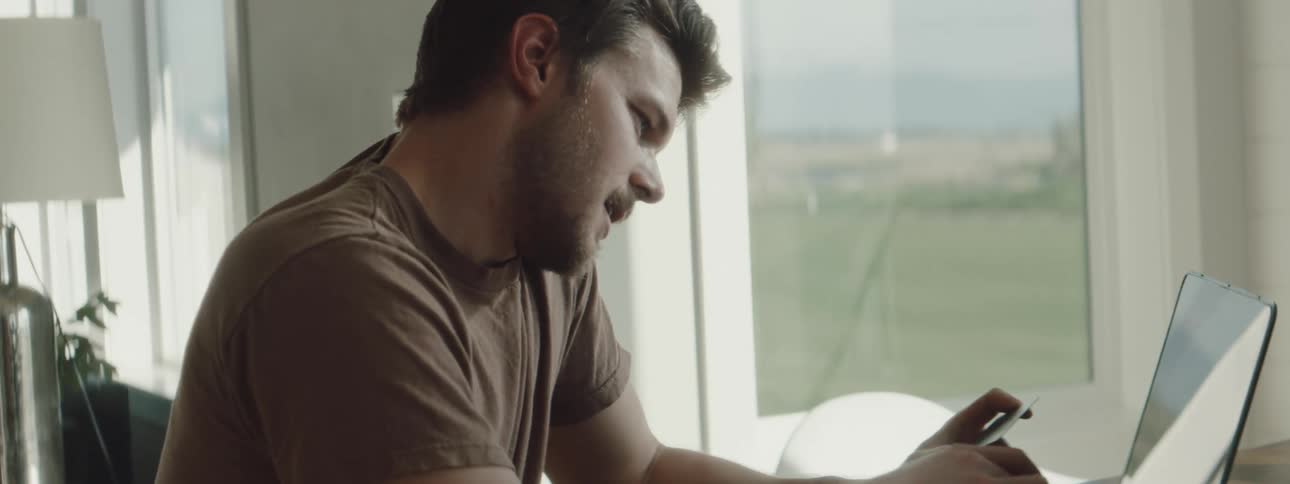 A man sitting at a table typing on a laptop with a lamp and a window in the background