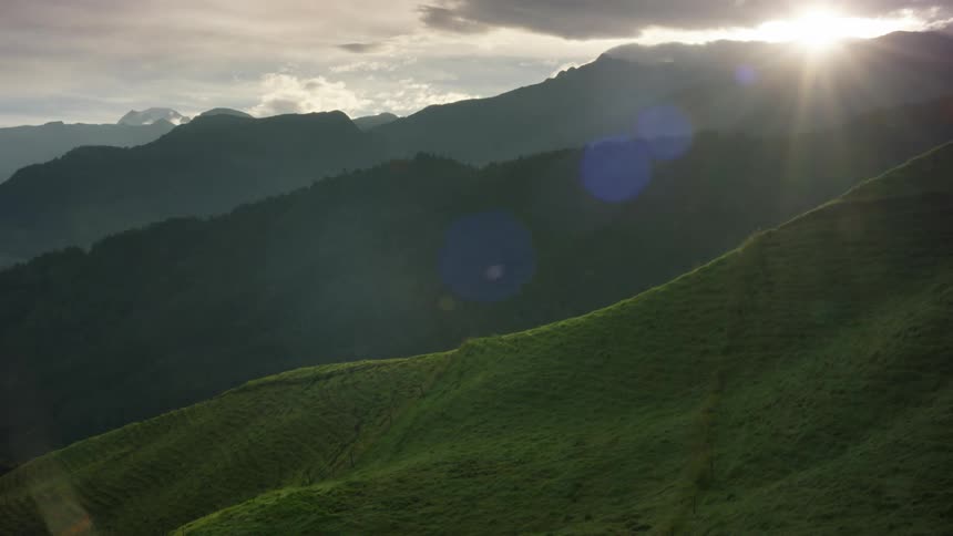 Grass covered hills and mountains under a cloud filled sky with the sun shining above a mountain