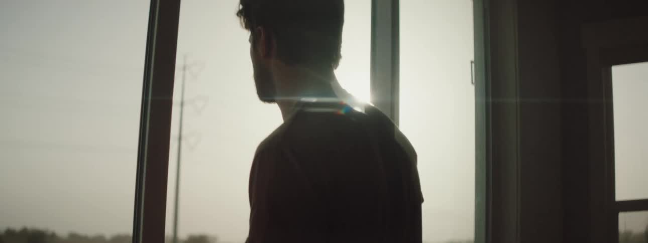 Man with a beard a mustache and a brown t shirt looks out the window as the sun shines brightly