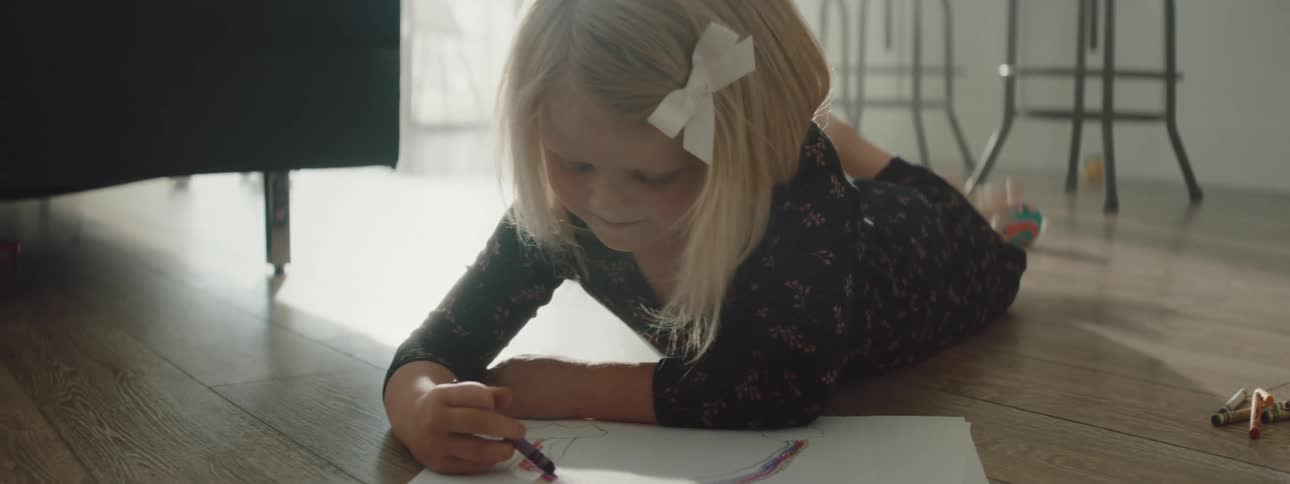 Little blonde girl drawing with crayons on a piece of paper on the living room floor