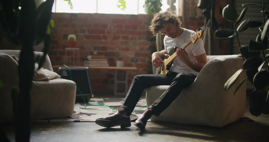 A man sitting on a couch playing a guitar with a wall and window in the background