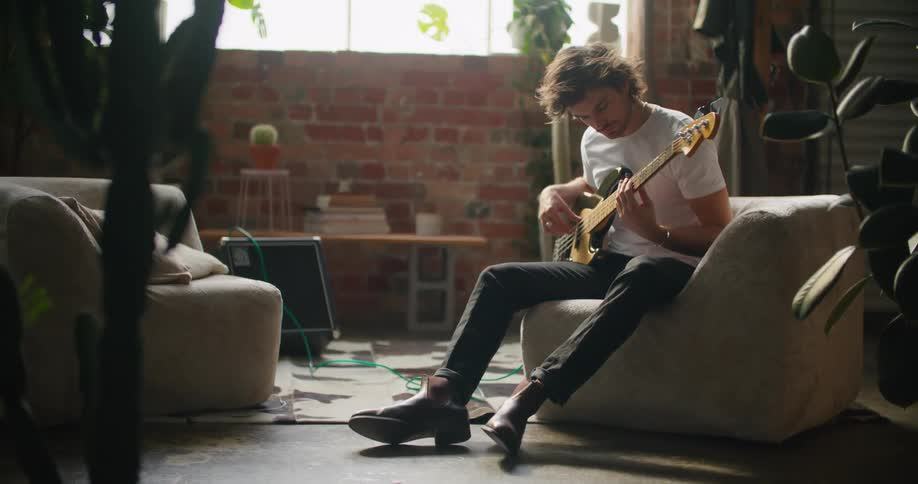 A man sitting on a couch in a room playing a guitar with a wall and window in the background