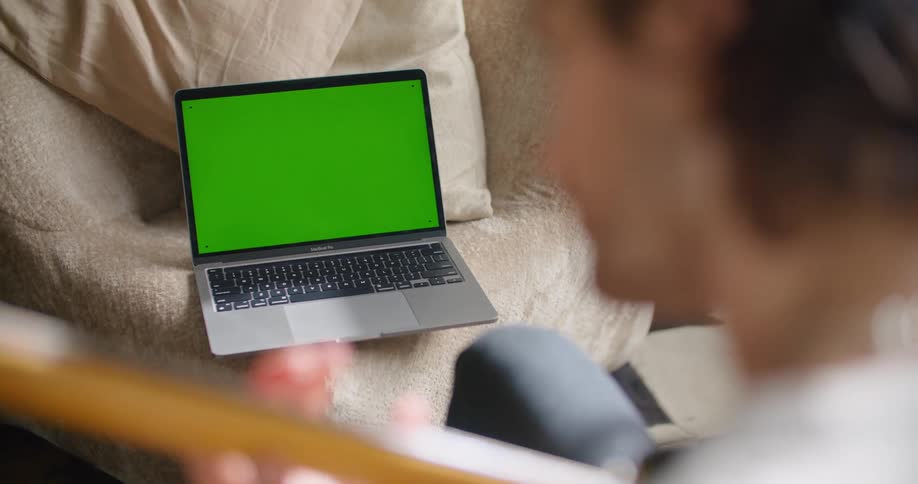 Man with a sweater and jeans plays guitar and looks at laptop with green screen