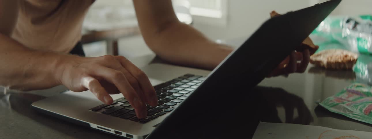 A man in a room typing on a laptop and talking on a smart phone with a wall and window in the background